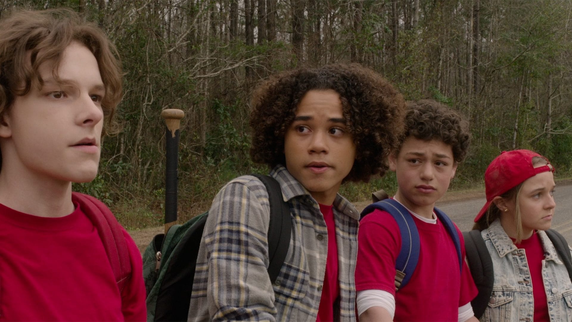 Afbeelding van vier kinderen, allemaal met een rood accent in hun kleding, die wat terughoudend en twijfelend kijken. Ze staan op een weg, met op de achtergrond een bosrand.