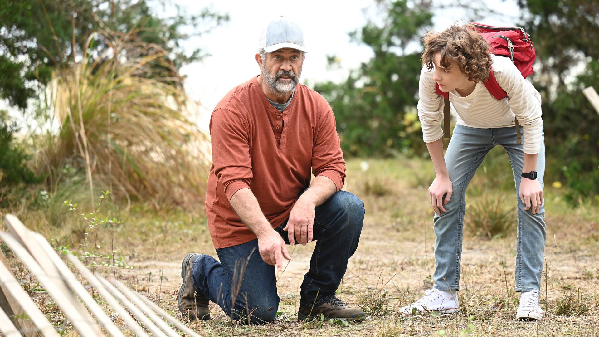 Afbeelding van Mel Gibson die op 1 knie gehurkt een verdachte situatie bekijkt, terwijl een ander kind met zijn handen op de knieën geïnteresseerd meekijkt. 