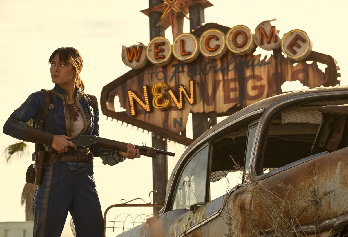 Afbeelding van een vrouw met een shotgun die langs een zwaar beschadigd Las Vegas welkomstbord loopt waar nu New Vegas op staat.