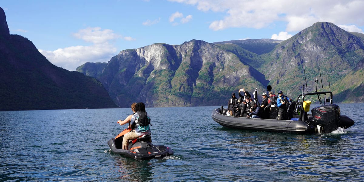 Afbeelding van een boot met mensen en een waterscooter waar twee mensen op zitten op een meer, omringd door bergen