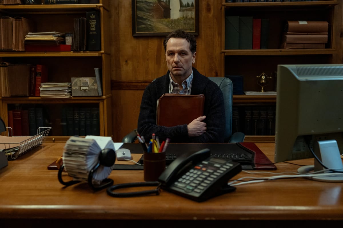 Een afbeelding van een man die aan een bureau zit met een map tegen zich aangedrukt. Hij kijkt niet blij.