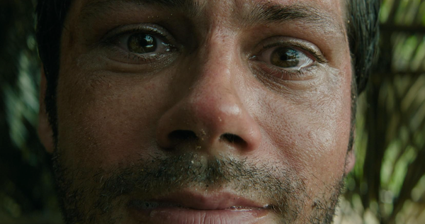 Op de afbeelding zie je een close-up van een man met betraand gezicht in een jungle omgeving, waarschijnlijk in shock.